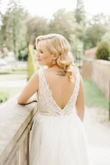  Blondynka w koronkowej sukni ślubnej z odkrytymi plecami, stojąca na tarasie z widokiem na zielony ogród. 