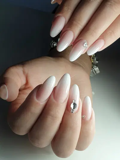  Elegancki manicure typu ombre w odcieniach nude z ozdobnymi cyrkoniami na długich paznokciach szpiczastego kształtu. 