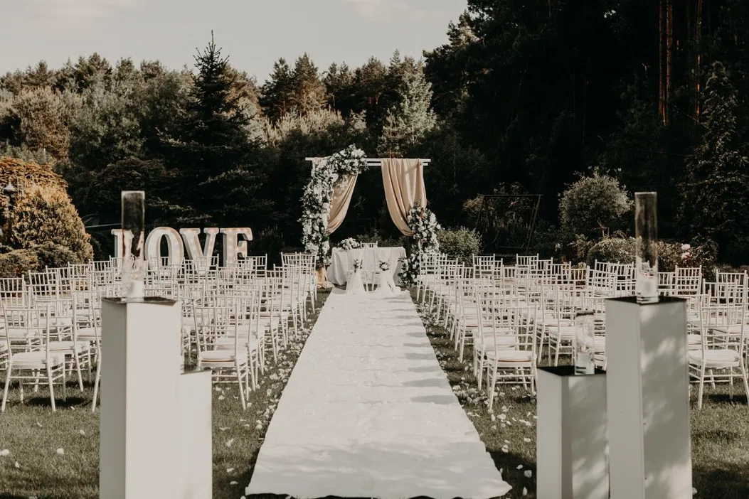 Plenerowa ceremonia ślubna z elegancką dekoracją kwiatową i białymi krzesłami ustawionymi w rzędach, napis LOVE w tle, zielony ogród w otoczeniu natury.