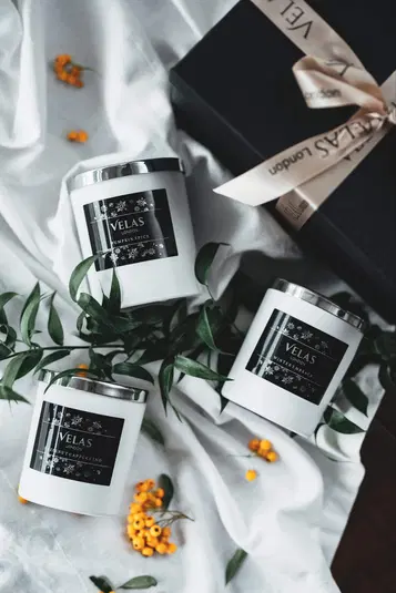  Białe świece zapachowe Velas London z etykietami Pumpkin Spice, Hazelnut Cappuccino i Winter Embrace, ustawione na białym tle z zielonymi liśćmi i żółtymi jagodami, obok eleganckiego czarnego pudełka z kremową kokardą. 