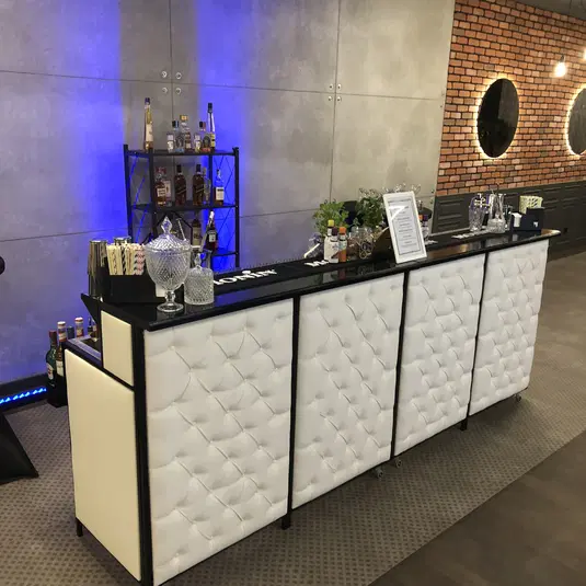  Luksusowy bar koktajlowy z białym, pikowanym frontem w nowoczesnym wnętrzu, ozdobiony butelkami alkoholi i elegancką zastawą, z widocznym oświetleniem led. 