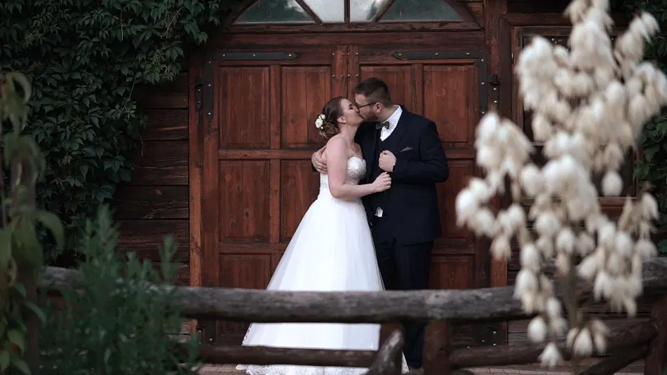  Para młoda w eleganckich strojach ślubnych całuje się przed drewnianymi drzwiami, fotografia ślubna w rustykalnym klimacie. 