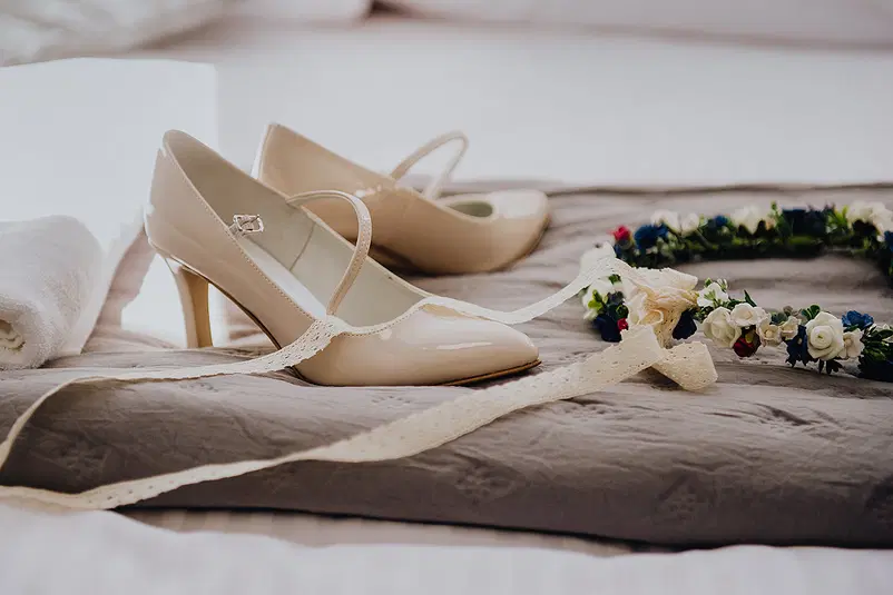  Beżowe buty na obcasie z koronką i kwiatowym wiankiem na tle szarej narzuty. 