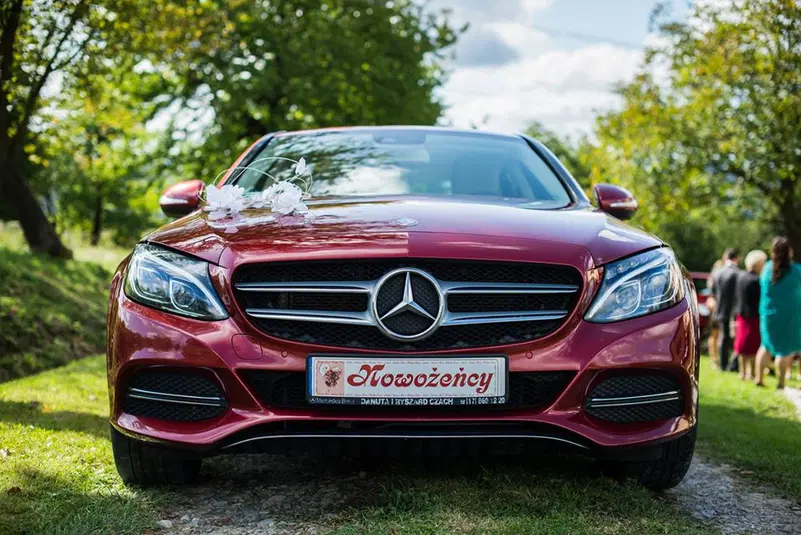  Czerwony Mercedes z dekoracją ślubną, widoczny z przodu, stojący na trawiastej alejce w słoneczny dzień. 