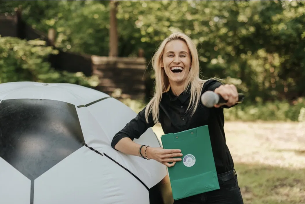 Kobieta trzymająca mikrofon, opierająca się o dużą inflatable piłkę nożną, z zieloną teczką i uśmiechem na twarzy, w parku podczas słonecznego dnia.