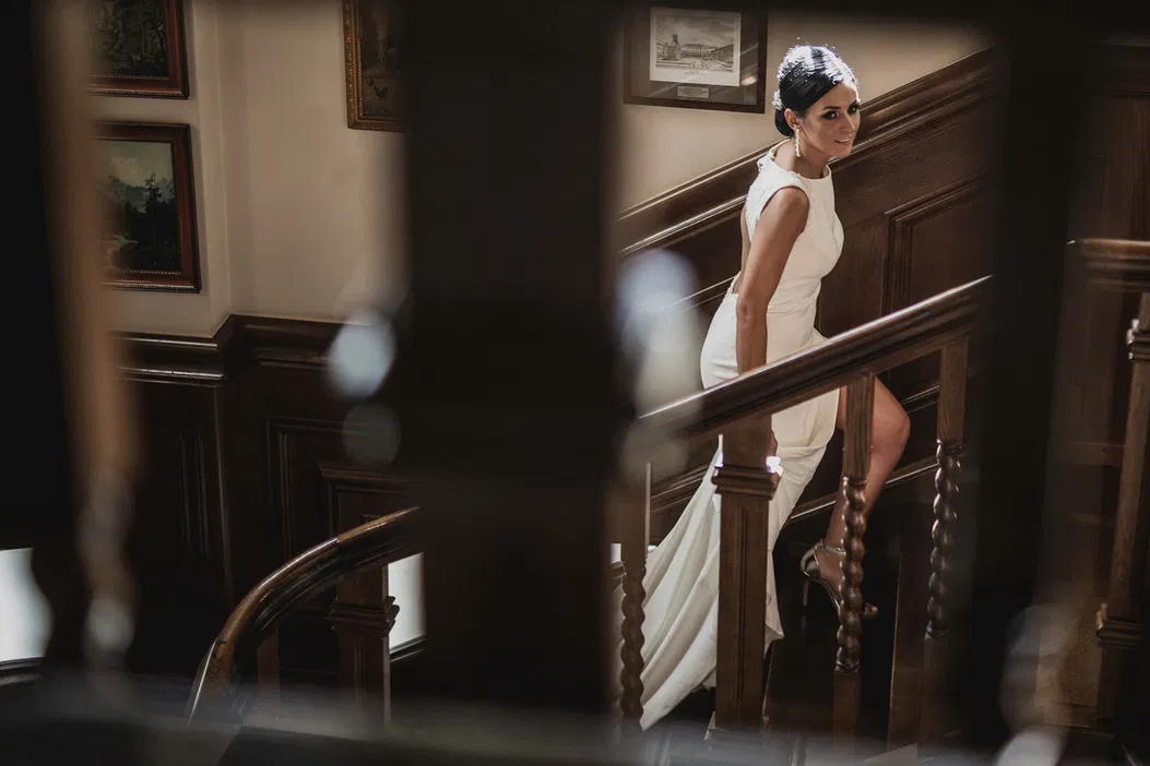 Kobieta w eleganckiej białej sukni idzie po drewnianych schodach w stylowym wnętrzu.