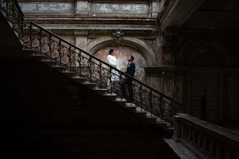  Stare schody w zabytkowym budynku z parą elegancko ubranych ludzi, kobieta w białym płaszczu i mężczyzna w niebieskim garniturze, stojących naprzeciw siebie. 