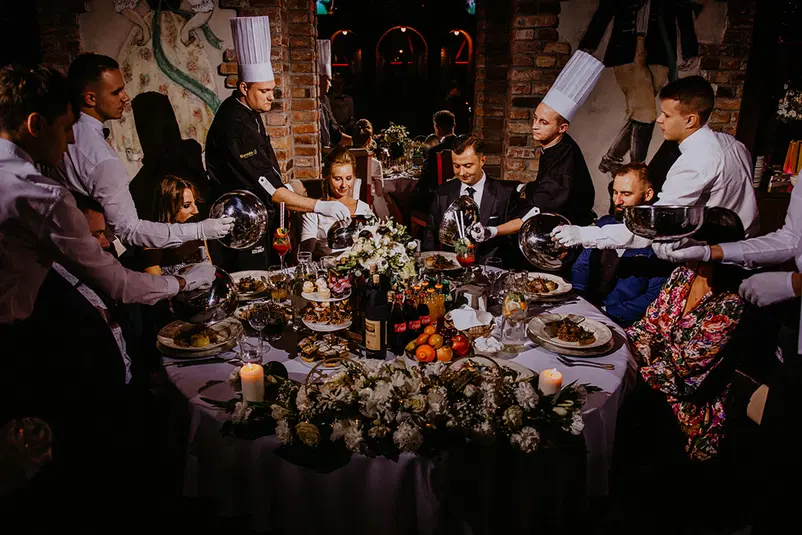  Luksusowa kolacja na przyjęciu weselnym z elegancko ubraną obsługą, udekorowanym stołem i różnorodnymi potrawami oraz napojami podawanymi gościom. 