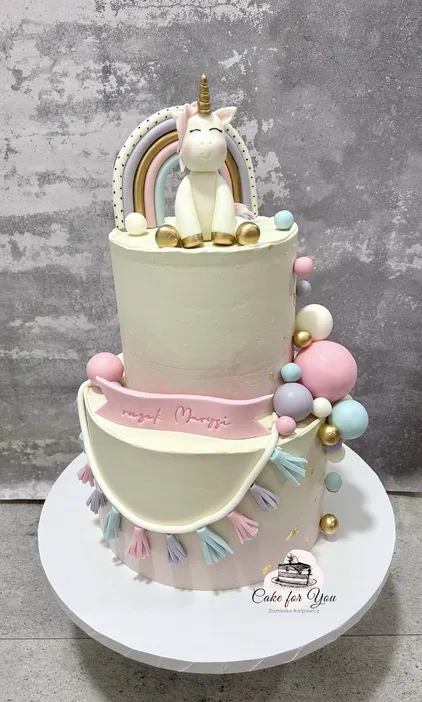 Dwupiętrowy tort urodzinowy z jednorożcem i tęczą, pastelowymi dekoracjami w różu, błękicie i złocie, ozdobiony napisem Roczek Marysi.