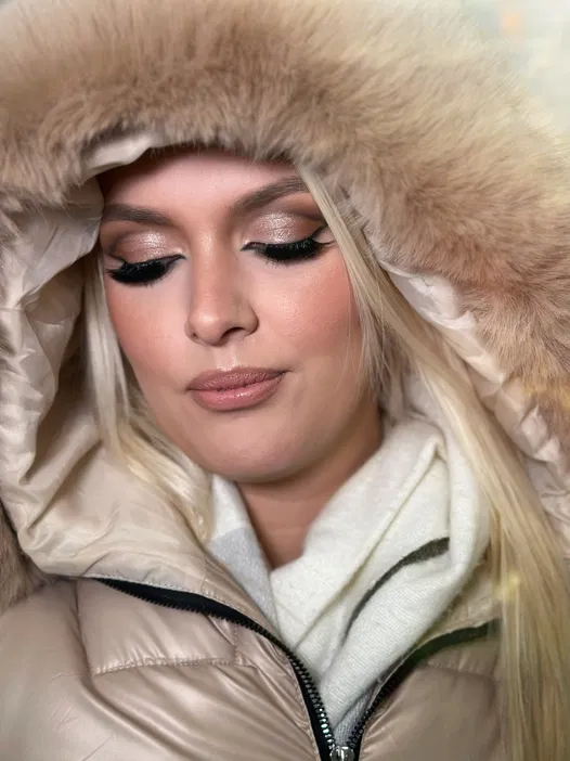 Blondynka w zimowej kurtce z kapturem wykończonym futrem, z profesjonalnym makijażem oczu, patrząca w dół.