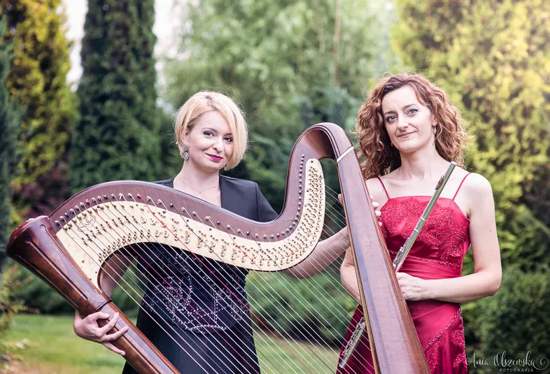  Dwie kobiety w eleganckich sukniach z harfą i fletnią na tle zieleni w ogrodzie. 