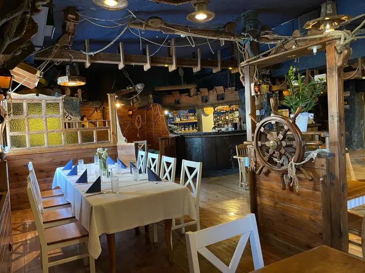  Wnętrze restauracji w stylu marynistycznym z drewnianymi dekoracjami, kapitańskim sterem i starannie nakrytym stołem. 