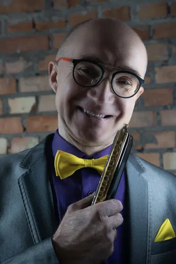  Mężczyzna z harmonicą w dłoni, w okularach, żółtej muszce i szarym garniturze, na tle ceglanej ściany. 
