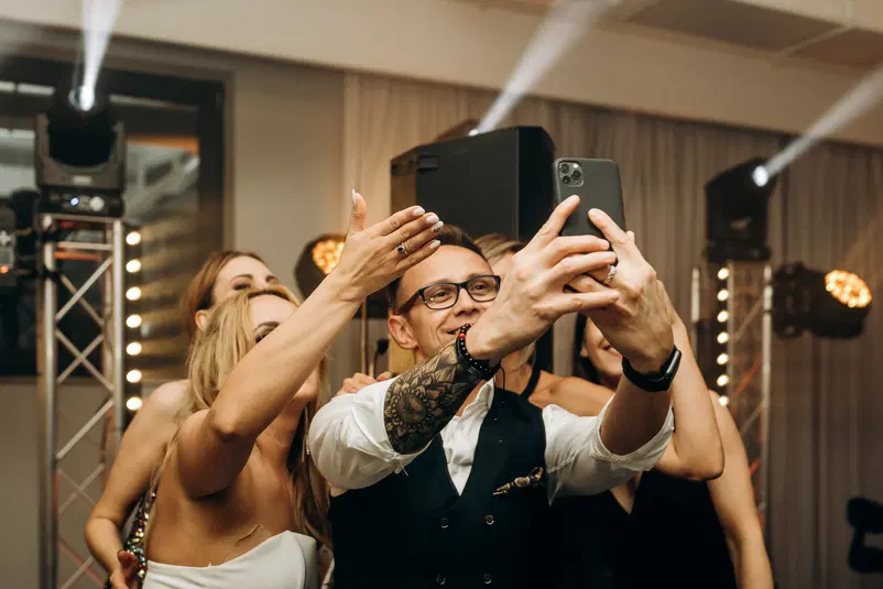  Grupa młodych ludzi robiąca selfie podczas imprezy z oświetleniem estradowym w tle, mężczyzna z tatuażem na ramieniu i zegarkiem na ręce, uśmiechnięci uczestnicy w eleganckich strojach. 