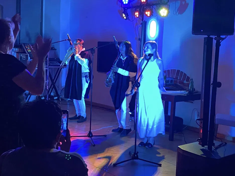 Trio muzyczne na scenie podczas koncertu, jeden z członków gra na fletni pana, inny na saksofonie; kolorowe oświetlenie i publiczność w tle, nocna atmosfera.