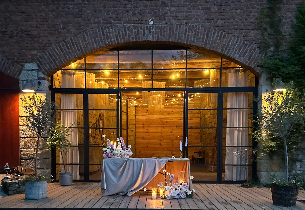 Romantyczna scena ślubna w rustykalnym stylu, dekoracje kwiatowe na drewnianym tarasie z oświetleniem świątecznym, szklane drzwi w stylu loftowym, ceremonia wieczorna.
