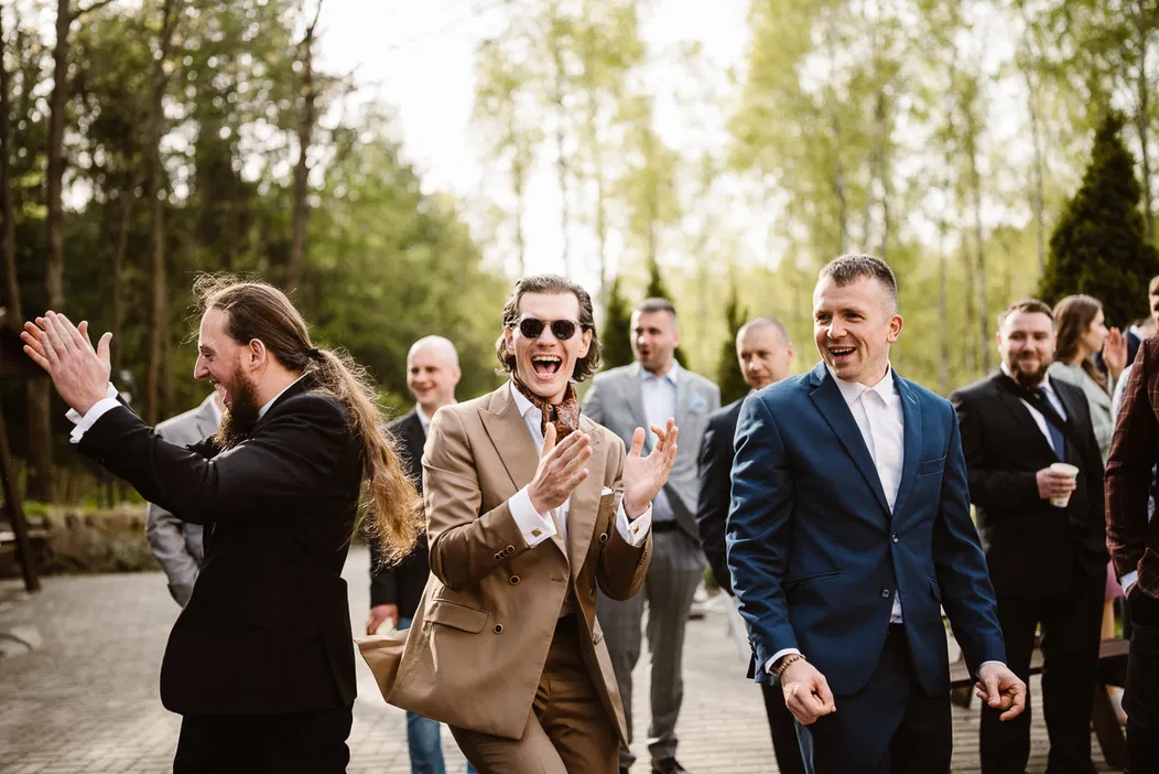 Grupa uśmiechniętych mężczyzn w eleganckich garniturach, uczestniczących w weselu na świeżym powietrzu.