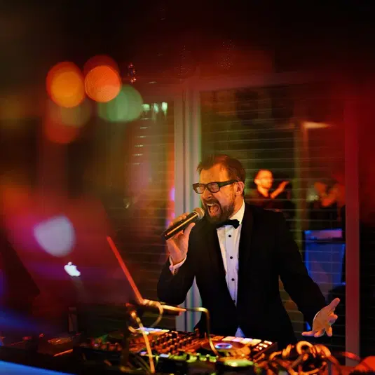  DJ śpiewający w eleganckim stroju podczas imprezy z kolorowymi światłami i sprzętem muzycznym w tle. 