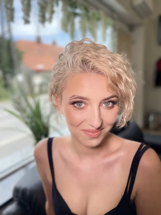 Nieznajoma kobieta z krótkimi, blond włosami, siedzi na sofie w jasnym pokoju z roślinami w tle, uśmiechając się delikatnie.