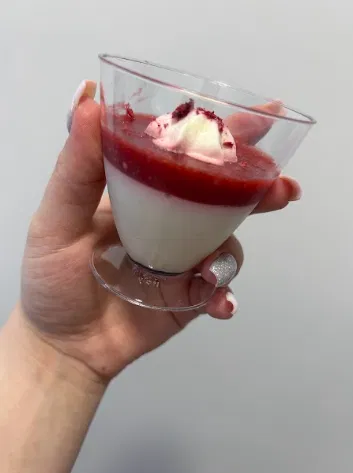 Ręka trzymająca deser panna cotta z sosem malinowym w przezroczystym kieliszku.