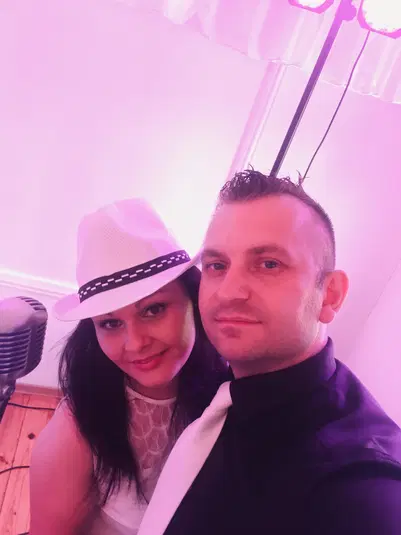  Bezpośredni kadr z imprezy, para elegancko ubrana, mężczyzna w czarnej koszuli z białym krawatem, kobieta w białym kapeluszu z czarną opaską, stoją obok mikrofonu, różowe oświetlenie w tle. 