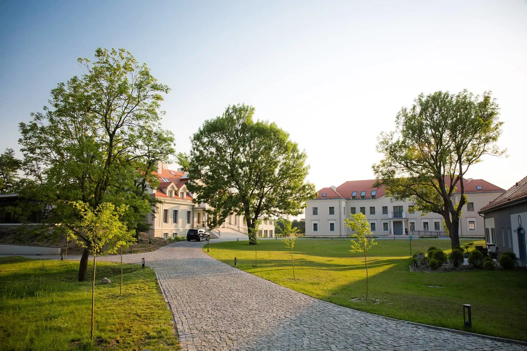 Zabytkowy pałac otoczony zielenią z brukowaną alejką i parkiem, letnie popołudnie.