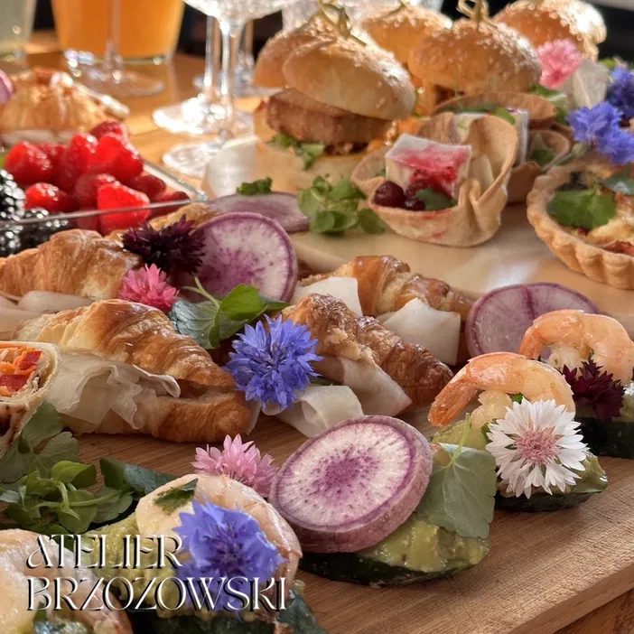 Rozmaite przysmaki na eleganckim przyjęciu: croissanty, owoce, burgery, tarty i dekoracyjne kwiaty, serwowane na drewnianym stole w Atelier Brzozowski.