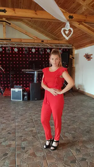  Kobieta w czerwonym kombinezonie na tle sceny z czerwonymi światłami LED, wnętrze sali z drewnianym sufitem. 