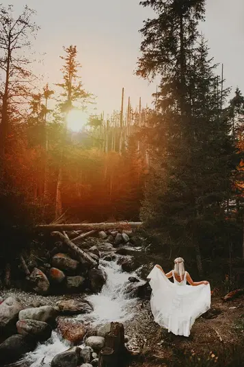  Panna młoda w białej sukni stojąca nad górskim potokiem w lesie o zachodzie słońca, romantyczna fotografia ślubna na łonie natury. 