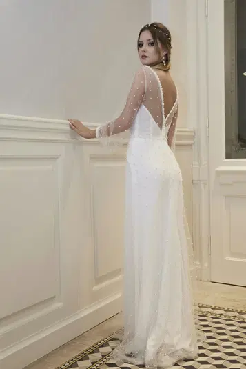  Kobieta w eleganckiej białej sukni ślubnej z koronkowymi rękawami, stojąca w eleganckim wnętrzu przy ścianie. 