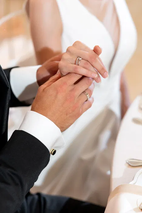 Bliskie ujęcie trzymającej się pary młodej w eleganckiej stylizacji ślubnej, z widocznymi obrączkami i suknią ślubną w tle.