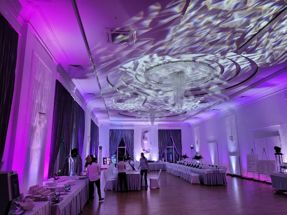 Elegancka sala bankietowa z nowoczesnym oświetleniem fioletowym, stoły biesiadne ustawione w zestawieniach, dekoracje świetlne tworzące wzory na suficie.