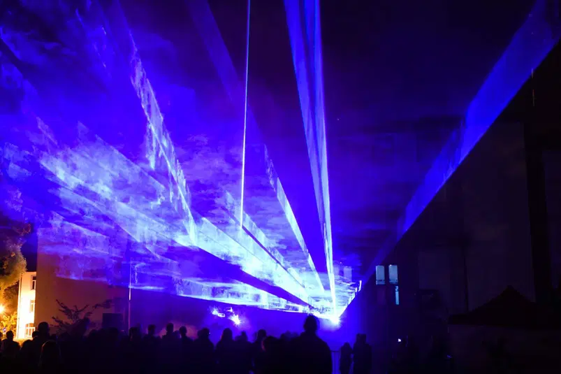  Widok naświetla laserowe na nocnym niebie na koncercie, z sylwetkami ludzi na pierwszym planie. 
