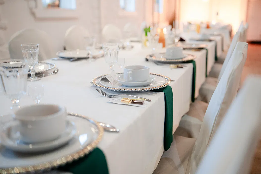 Elegancko nakryty stół na przyjęcie z białymi obrusami, porcelanowymi talerzami, kryształowymi szklankami i zielonymi serwetkami.