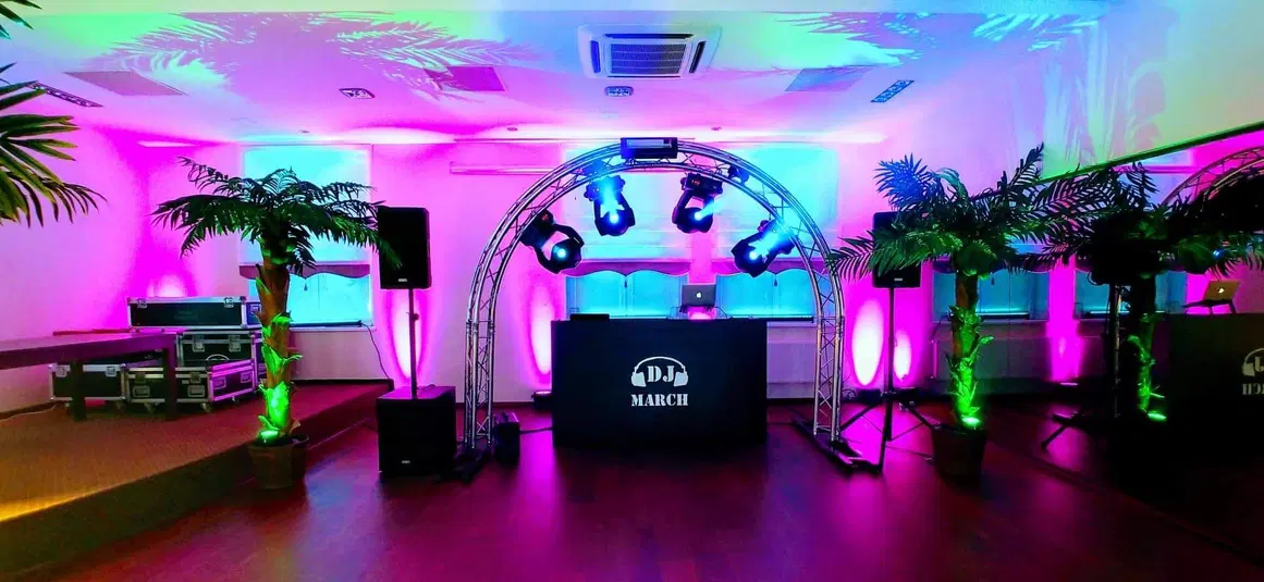  Kolorowe oświetlenie DJ na imprezie; scena z podestem, reflektorami i efektami świetlnymi; dekoracje roślinne; sprzęt muzyczny i nagłośnieniowy; klubowa atmosfera. 
