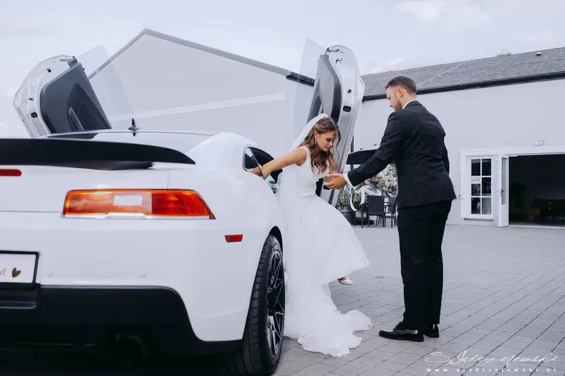  Para młoda przy białym sportowym samochodzie podczas ślubu, panna młoda w białej sukni opuszcza auto, pan młody w garniturze pomaga jej wysiąść, elegancka ceremonia ślubna na zewnątrz 