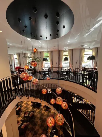  Dwupoziomowe wnętrze restauracji z nowoczesnym żyrandolem, okrągłymi, pomarańczowymi lampami wiszącymi z sufitu oraz eleganckimi krzesłami i stołami z białymi obrusami. 