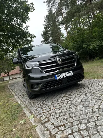  Czarny van Renault na wyłożonej kostką drodze, otoczony zielonymi drzewami. 