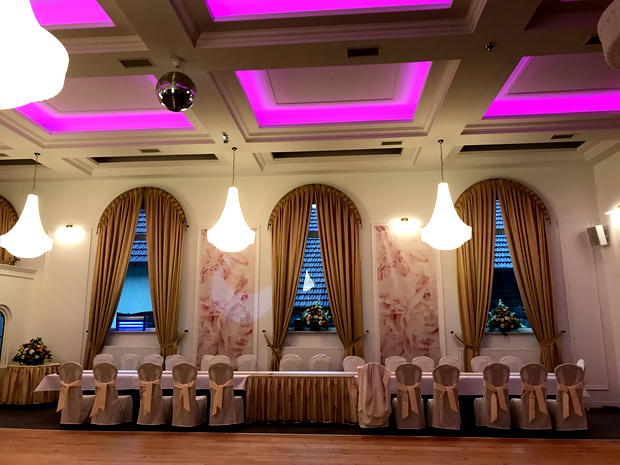  Elegancka sala bankietowa z podświetlanym sufitem i klasycznym wystrojem, długim stołem udekorowanym białymi pokrowcami na krzesła, eleganckimi zasłonami w oknach i dyskretnym oświetleniem. 