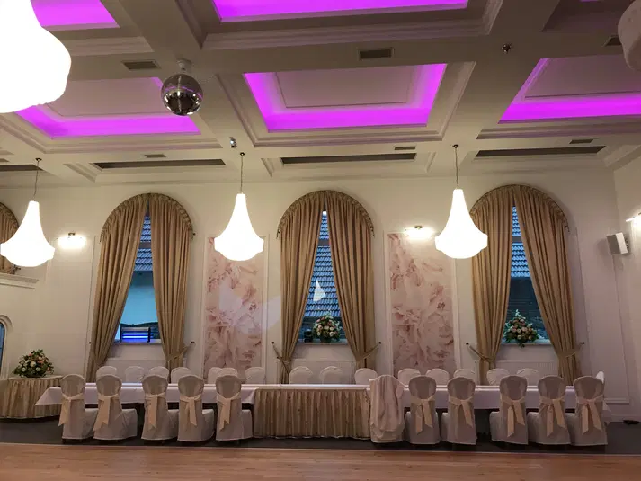  Elegancka sala bankietowa z podświetlanym sufitem i klasycznym wystrojem, długim stołem udekorowanym białymi pokrowcami na krzesła, eleganckimi zasłonami w oknach i dyskretnym oświetleniem. 