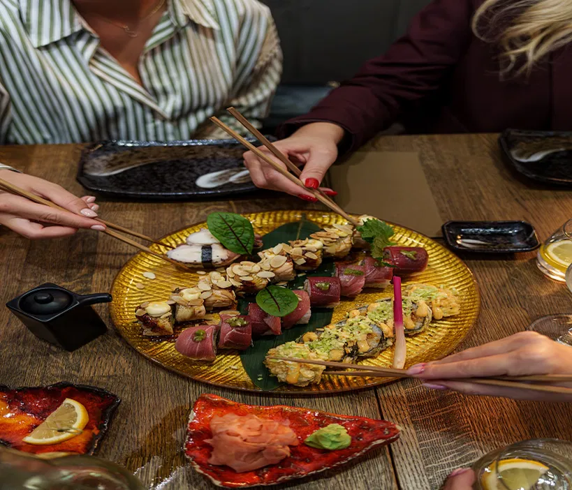 Ludzie jedzący sushi przy elegancko zastawionym stole w restauracji, wykwintne dania japońskie, wspólne posiłki, sztuka kulinarna, używanie pałeczek do jedzenia sushi, świeże składniki, spotkanie towarzyskie.