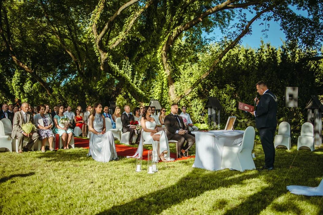 Ceremonia ślubna na świeżym powietrzu w otoczeniu natury, para młoda i goście siedzący na zielonej trawie pod drzewami, słoneczny dzień