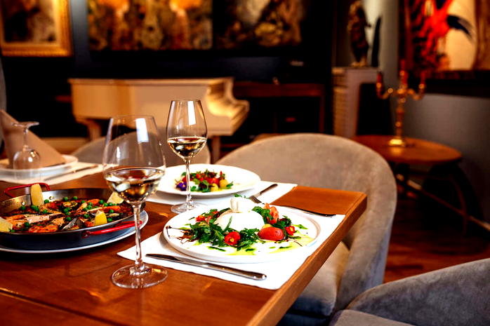  Elegancka restauracja z daniem z burraty i warzywami, kieliszki białego wina na stole, stylowe tło z pianinem i dekoracjami, kulinarne doświadczenie. 