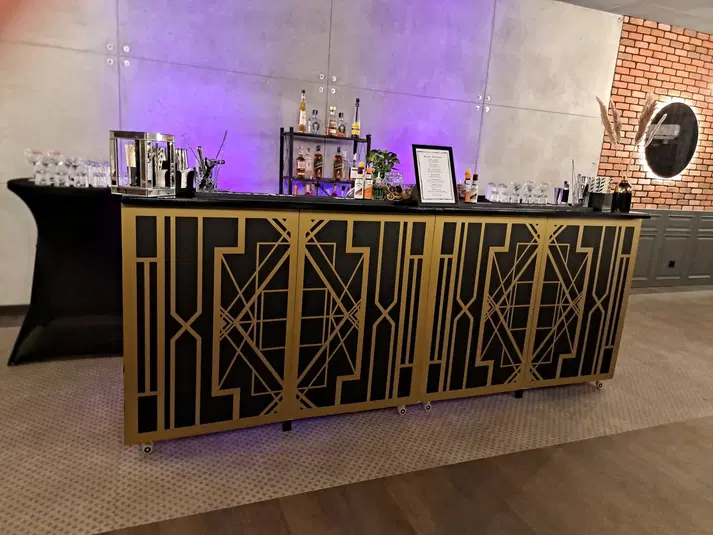  Stylowy bar Art Deco z ozdobionymi elementami geometrycznymi, wyposażony w alkohole i akcesoria barmańskie, na tle nowoczesnego wnętrza z ceglaną i betonową ścianą. 