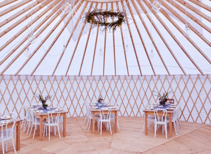  Yurt-style event venue interior with wooden tables, white chairs, floral centerpieces, and decorative ceiling wreath. 