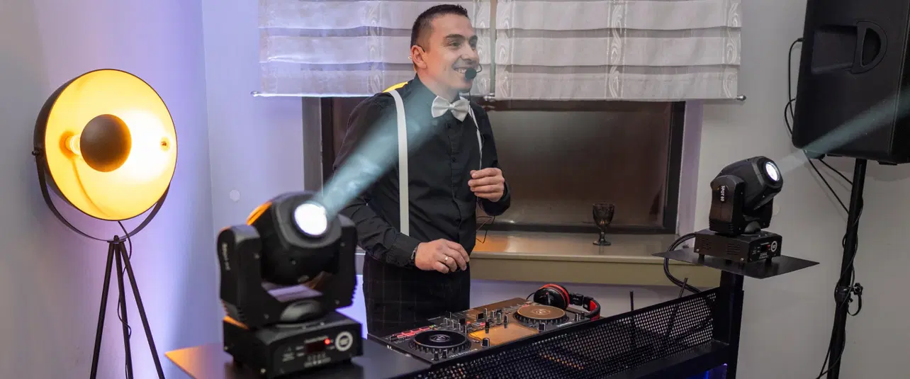  DJ na imprezie, konsola DJ-ska z oświetleniem, osoba z mikrofonem, profesjonalne nagłośnienie, elegancki strój, nowoczesne efekty świetlne. 