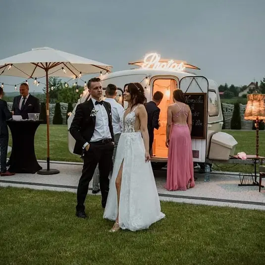 Para młoda w eleganckich strojach na weselu przed fotobudką na świeżym powietrzu, wieczorna atmosfera z dekoracyjnymi lampkami i gośćmi w tle. 