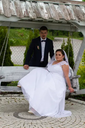  Para młoda w sesji ślubnej, pan młody w eleganckim garniturze, panna młoda w białej sukni na drewnianej huśtawce w ogrodzie. 
