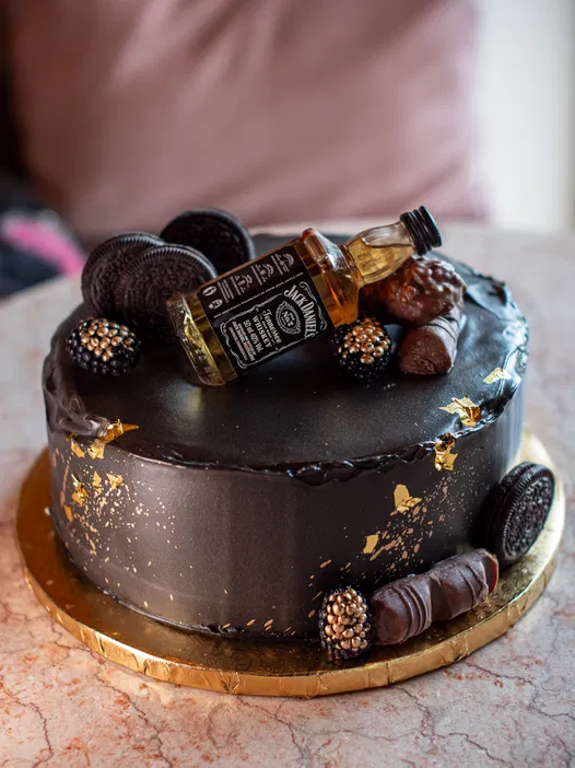 Czekoladowy tort zdobiony złotymi płatkami, miniaturową butelką whiskey, ciasteczkami Oreo i pralinkami na złotej podstawce.
