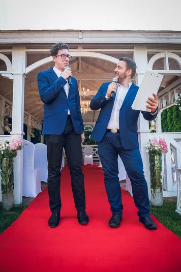  Dwóch mężczyzn w garniturach prowadzących imprezę na czerwonym dywanie na tle altany. 
